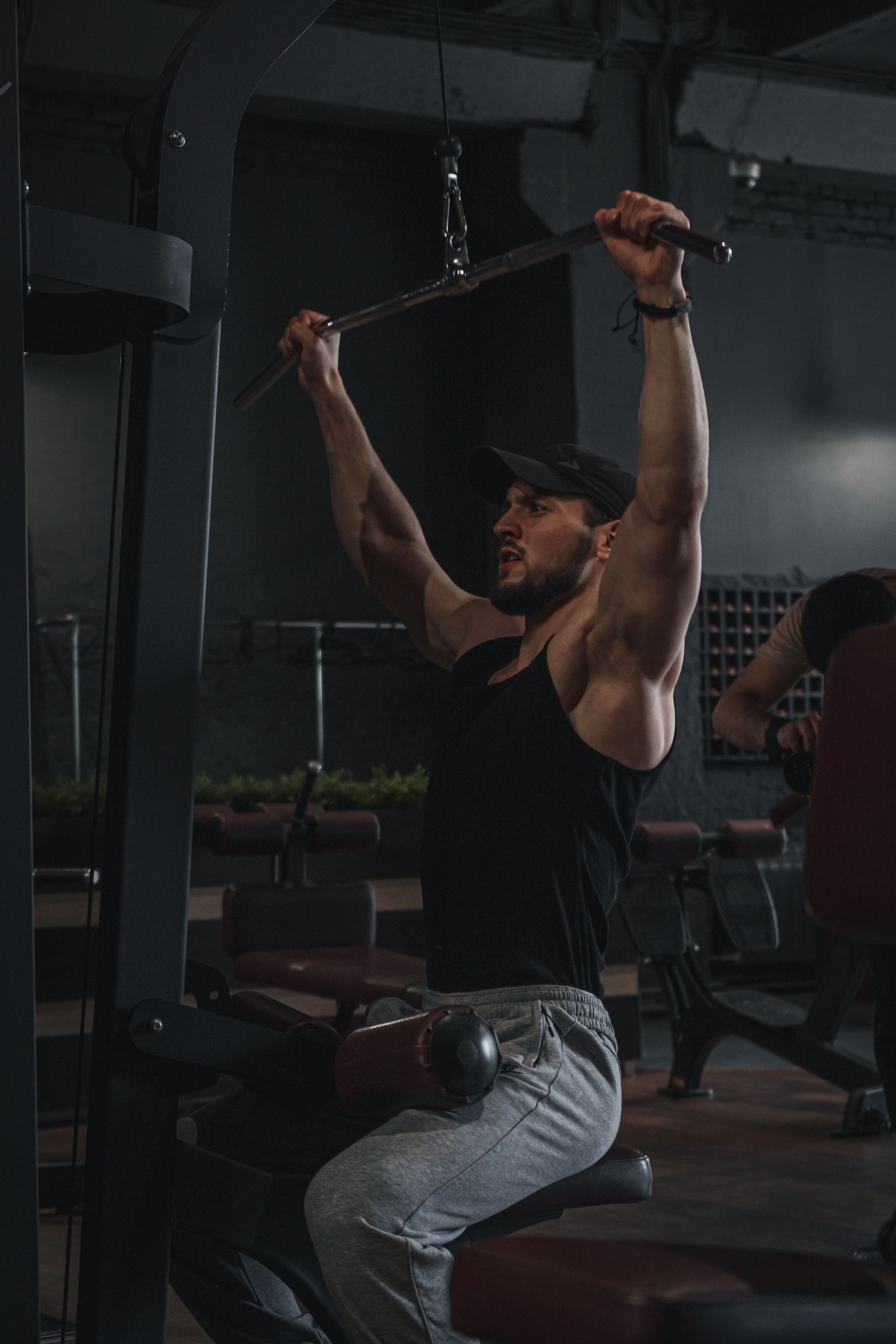 A man training alone on a lat pulldown in a dark gym, physique carved out of shadow