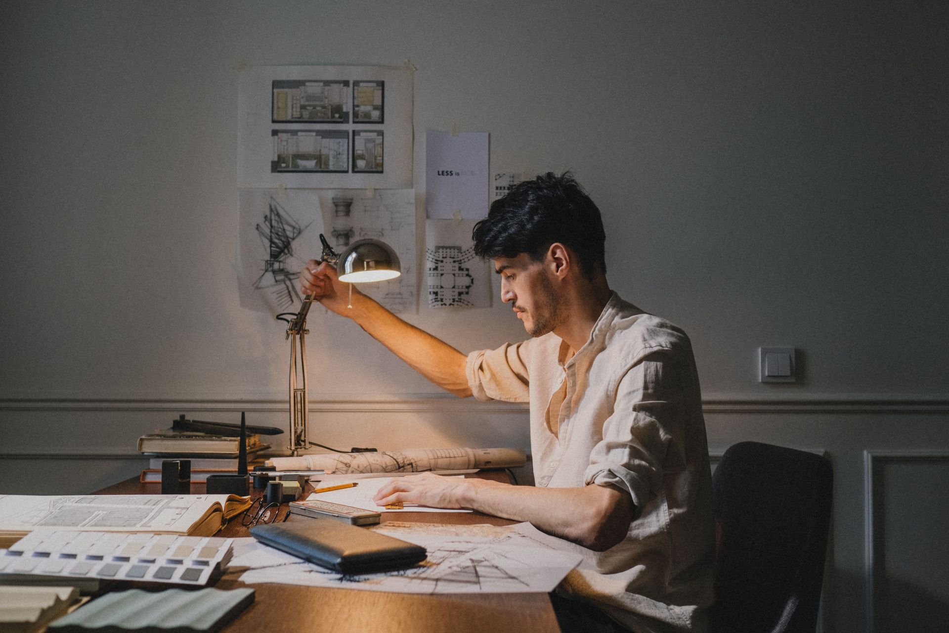 A man working through plans and sketches at a desk under one warm lamp in a dim room