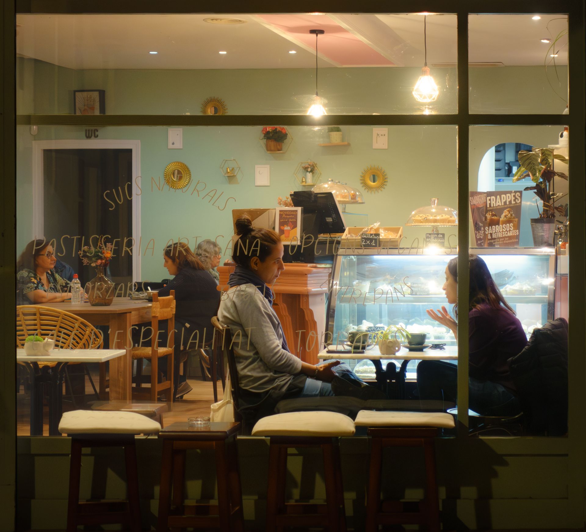 Two people deep in conversation in a warmly lit room at night, seen through a window