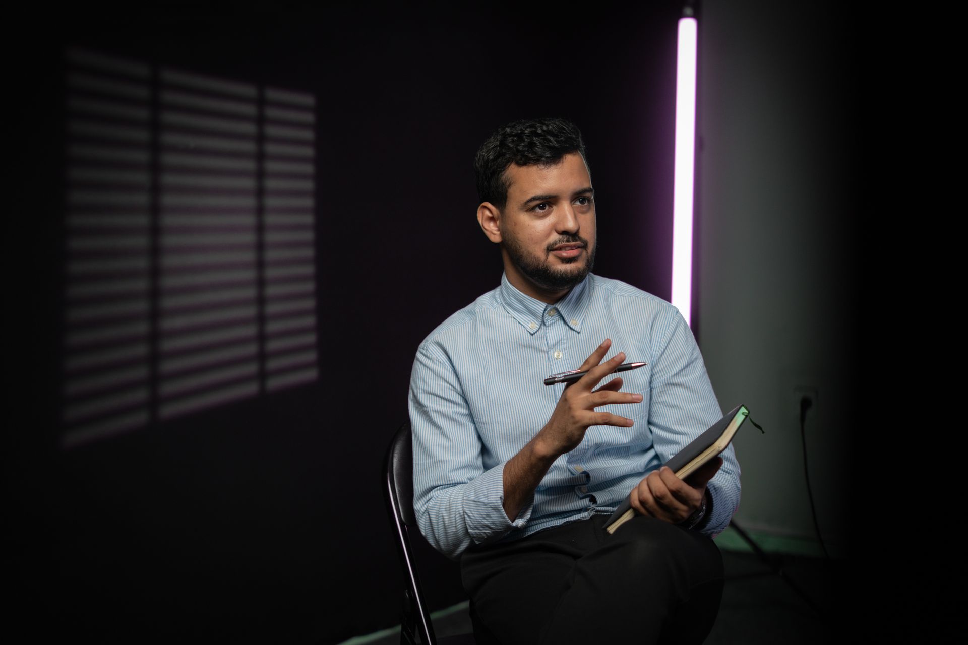 A man holding a notebook mid-conversation in a shadowed room lit by one violet light