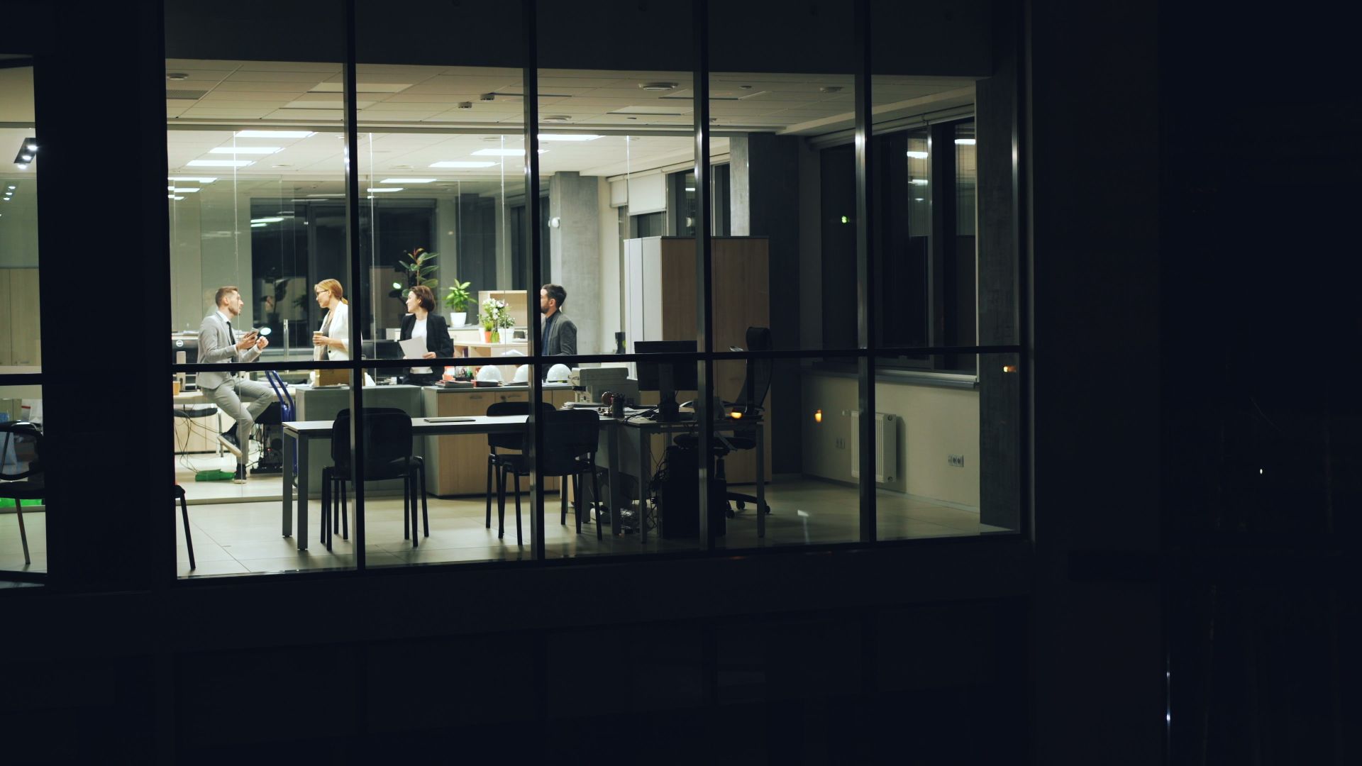 A team working late inside a glass-walled office, one warm room of activity in a dark building